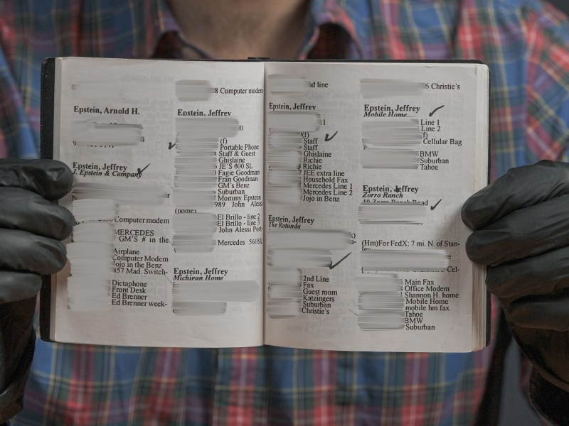 A person wearing black gloves holds open a small notebook displaying pages filled with handwritten and printed contact details, with several names and information redacted.