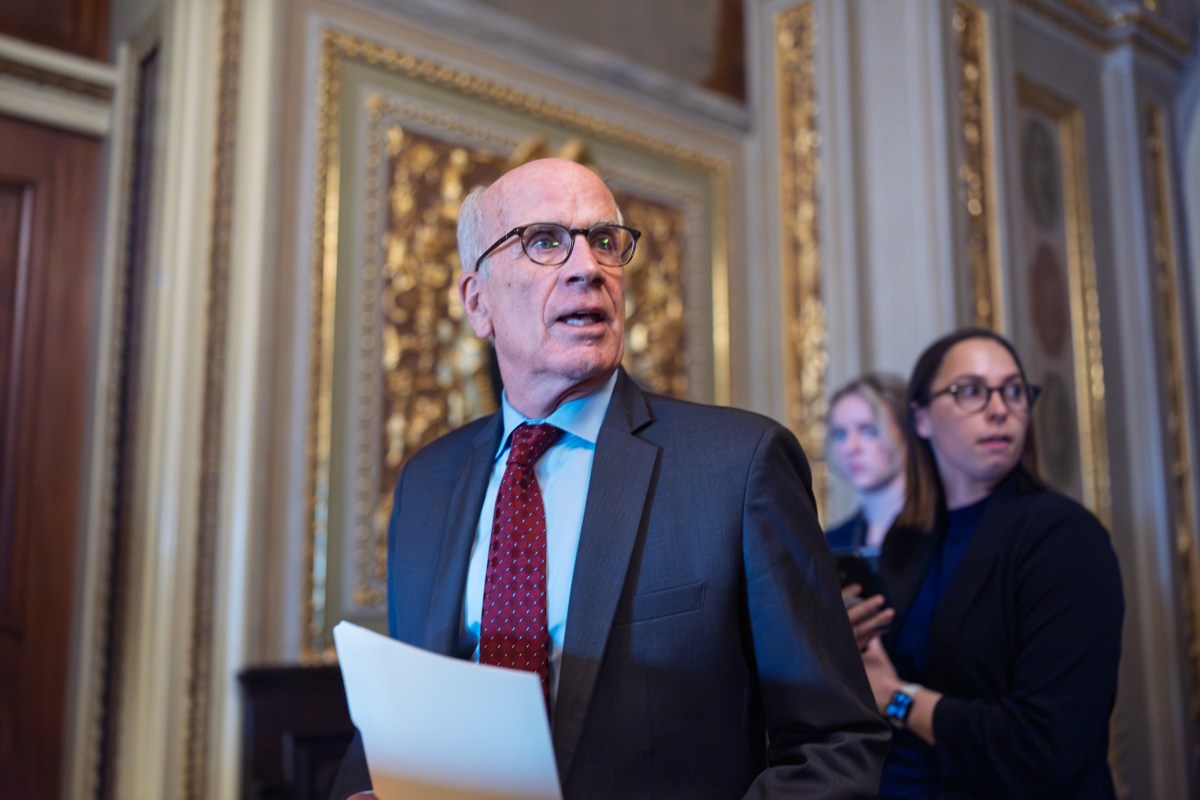 An older man in a suit holding papers stands in a richly decorated room, with two women in the background, one holding a phone.