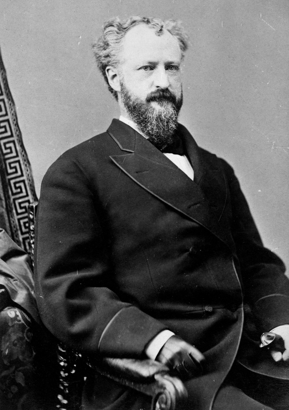 Black-and-white portrait of a seated man with a beard, wearing a formal suit and gloves, posing against a patterned backdrop.