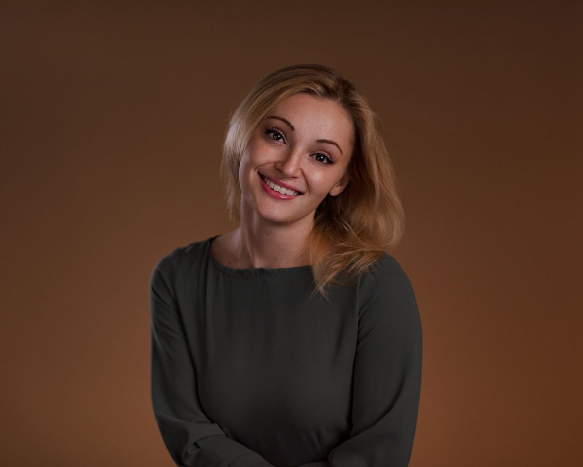 A woman with blonde hair, wearing a dark long-sleeve top, smiles at the camera in front of a plain brown background.