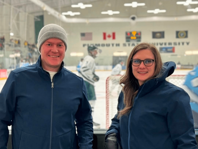 Two people in winter jackets stand in front of a hockey rink at Plymouth State University, with players and flags visible in the background.