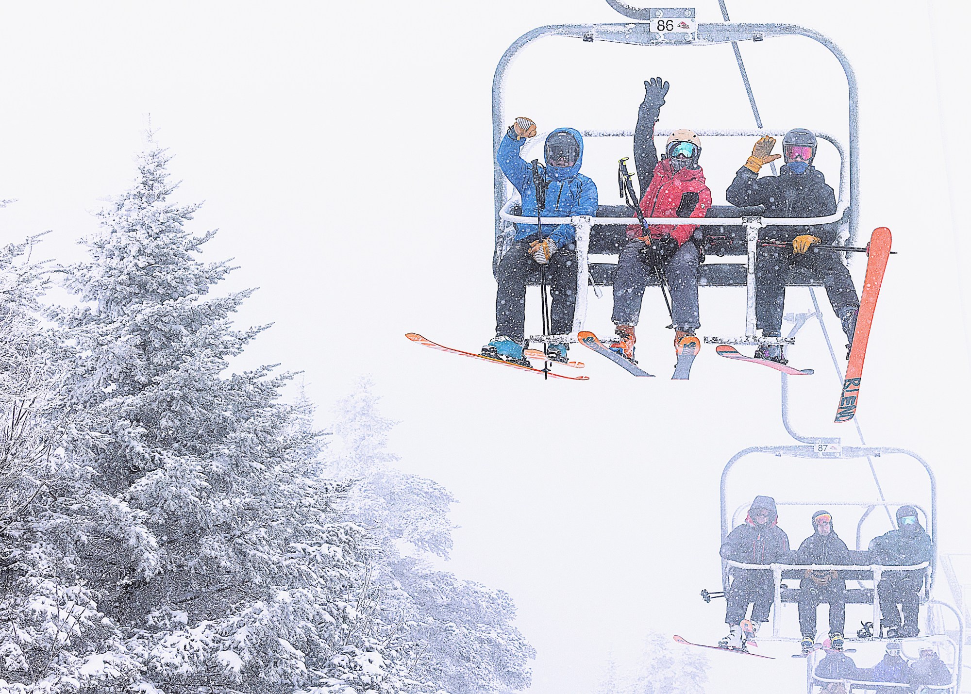 Four skiers sit on a snowy ski lift, waving at the camera, with snow-covered trees visible on the left side of the image.