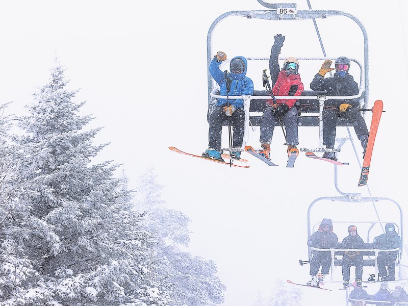 Four skiers sit on a snowy ski lift, waving at the camera, with snow-covered trees visible on the left side of the image.