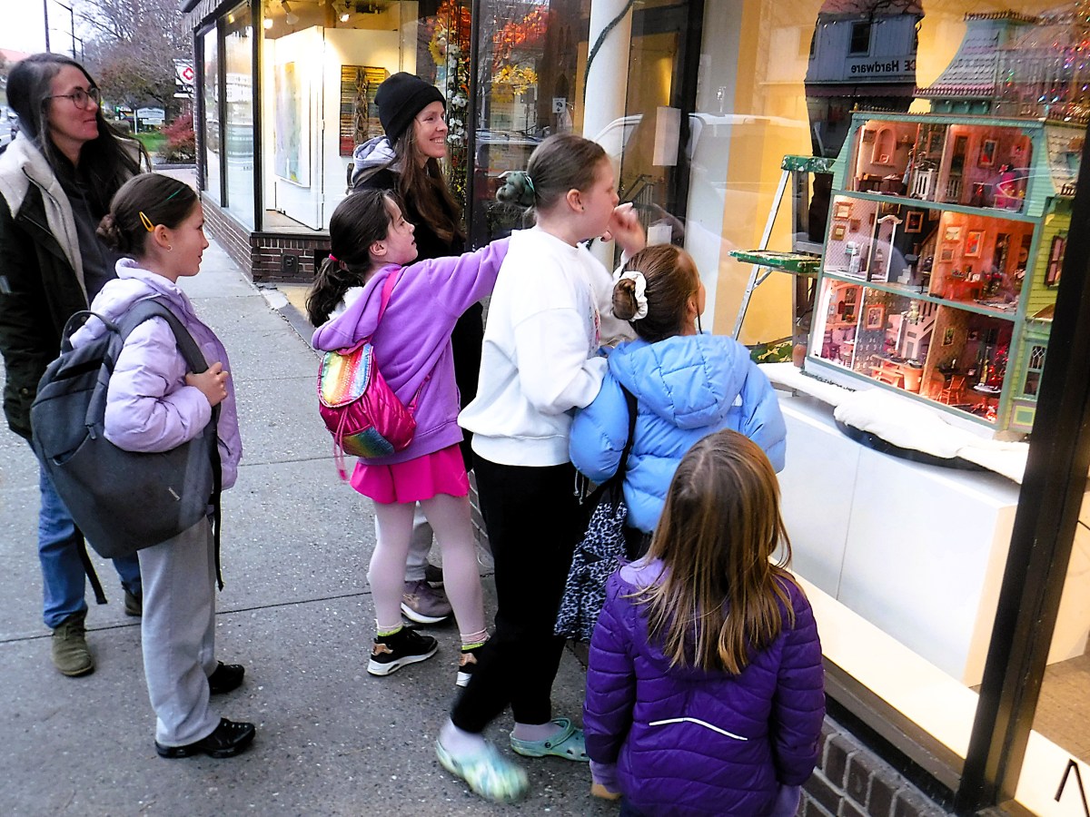 A group of children and two adults stand on a sidewalk, looking through a shop window at a large dollhouse display.