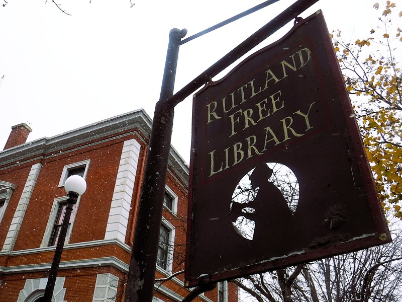 A close-up of a metal sign reading “Rutland Free Library” with a silhouette of a person reading, in front of a red-brick building on a cloudy, snowy day.