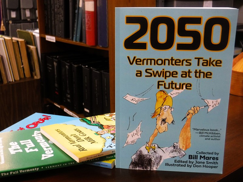 A book titled "2050: Vermonters Take a Swipe at the Future" stands upright on a library table, with two other books and shelves of files in the background.