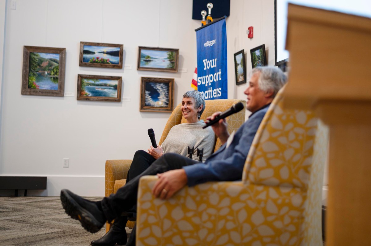 Two people sit in patterned chairs holding microphones, speaking at an indoor event. Framed landscape paintings hang on the wall, and a "Your support matters" banner is visible in the background.