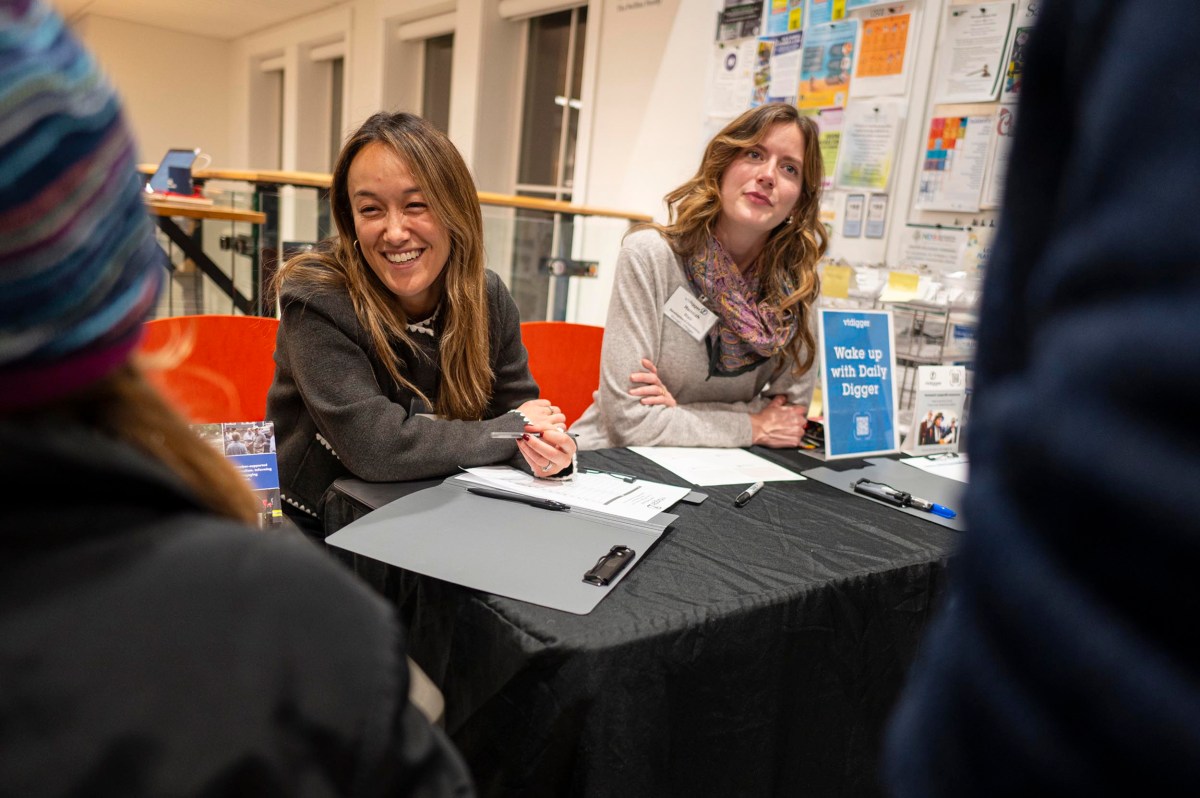 Two women sit at a table with clipboards and papers, engaging with people at an indoor event or registration area.