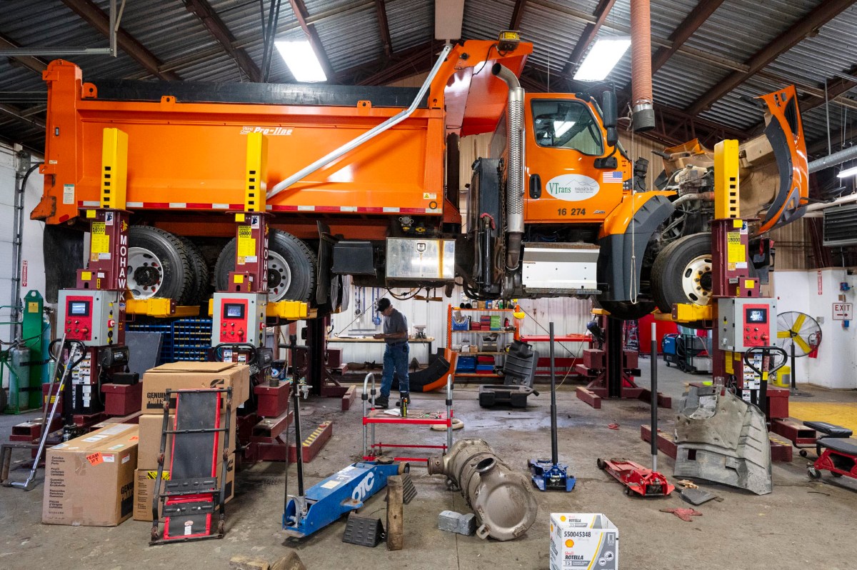 An orange dump truck is elevated on hydraulic lifts in a maintenance garage, surrounded by tools, parts, and equipment, while a person works underneath.