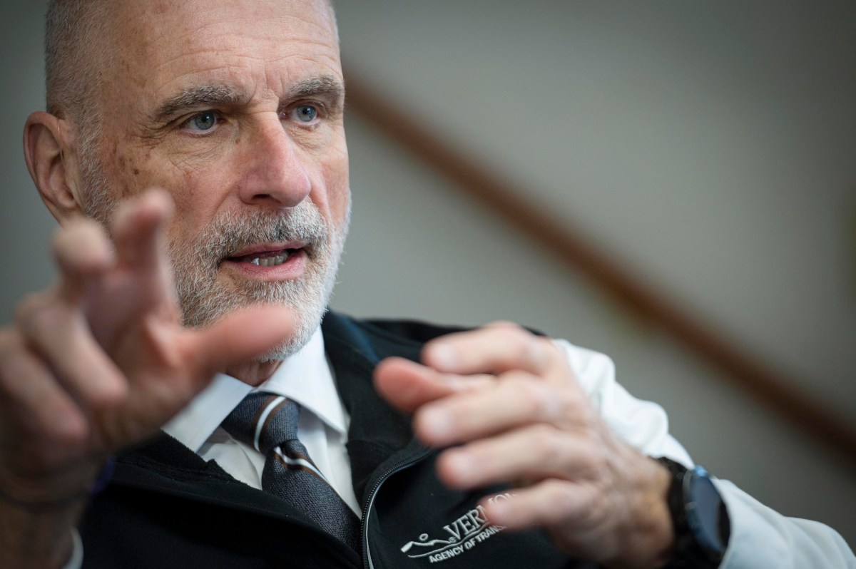 An older man with a trimmed gray beard gestures with his hands while speaking, wearing a tie, white shirt, and a vest that reads "Vermont" with a blurred background.