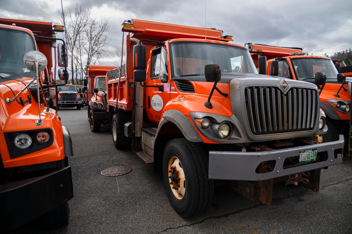 Several orange dump trucks are parked side by side in a lot under a cloudy sky.
