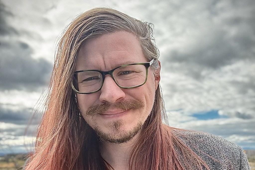 A person with long hair, glasses, and a mustache is standing outdoors under a cloudy sky, looking at the camera and smiling slightly.