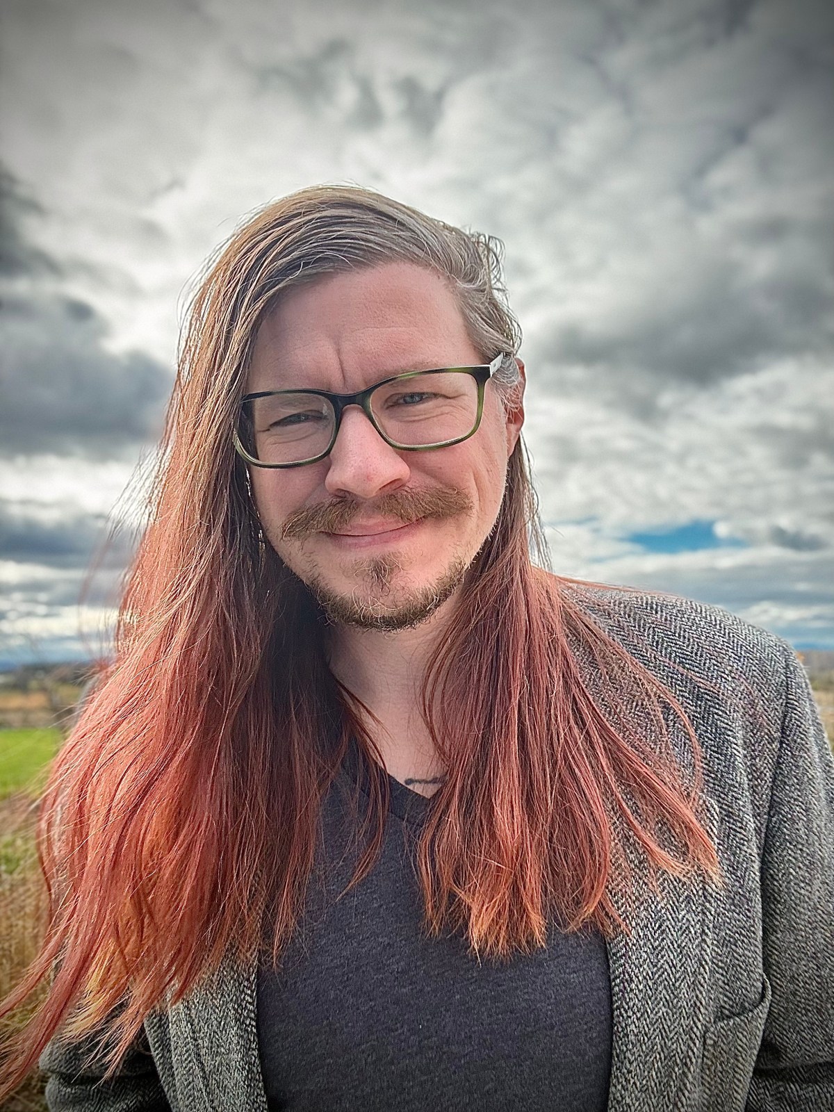 A person with long reddish-brown hair, glasses, and a beard stands outdoors wearing a gray jacket and dark shirt, with a cloudy sky and fields in the background.