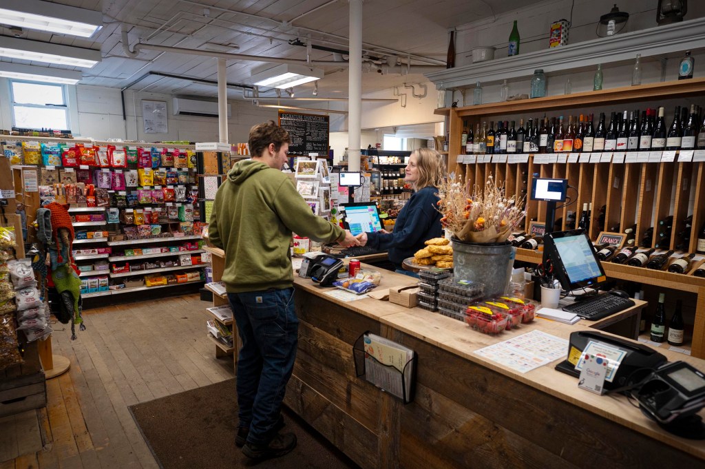 A customer stands at the counter speaking to a cashier in a small grocery store with shelves of snacks, drinks, and wine in the background.