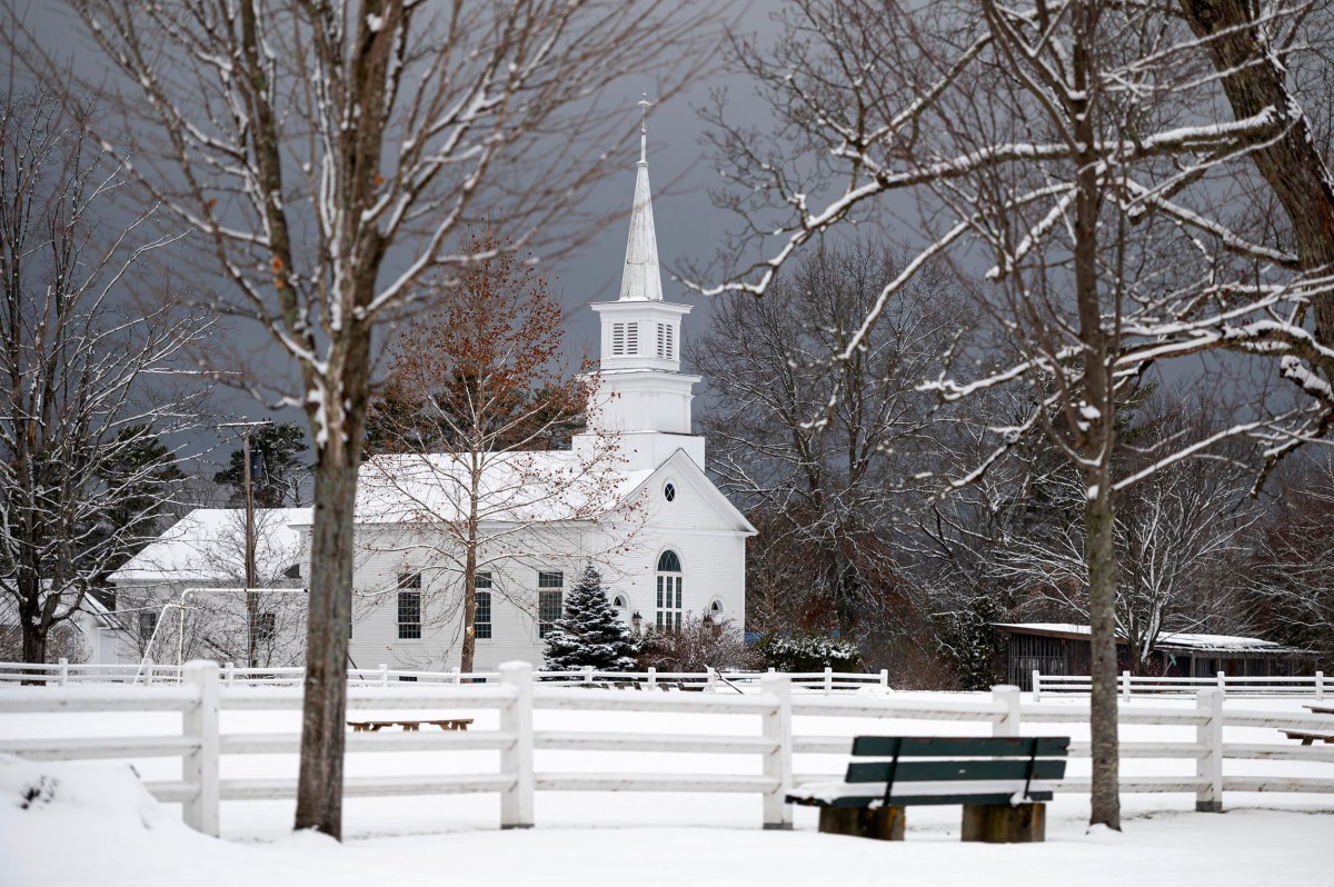 A white church with a steeple stands amid snow-covered trees and a white fence under an overcast sky; a green bench sits in the foreground.