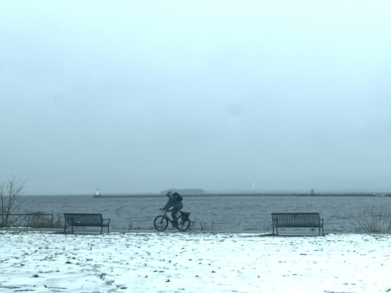 A person rides a bicycle on a snowy path near a body of water, with two empty benches and a cloudy sky in the background.
