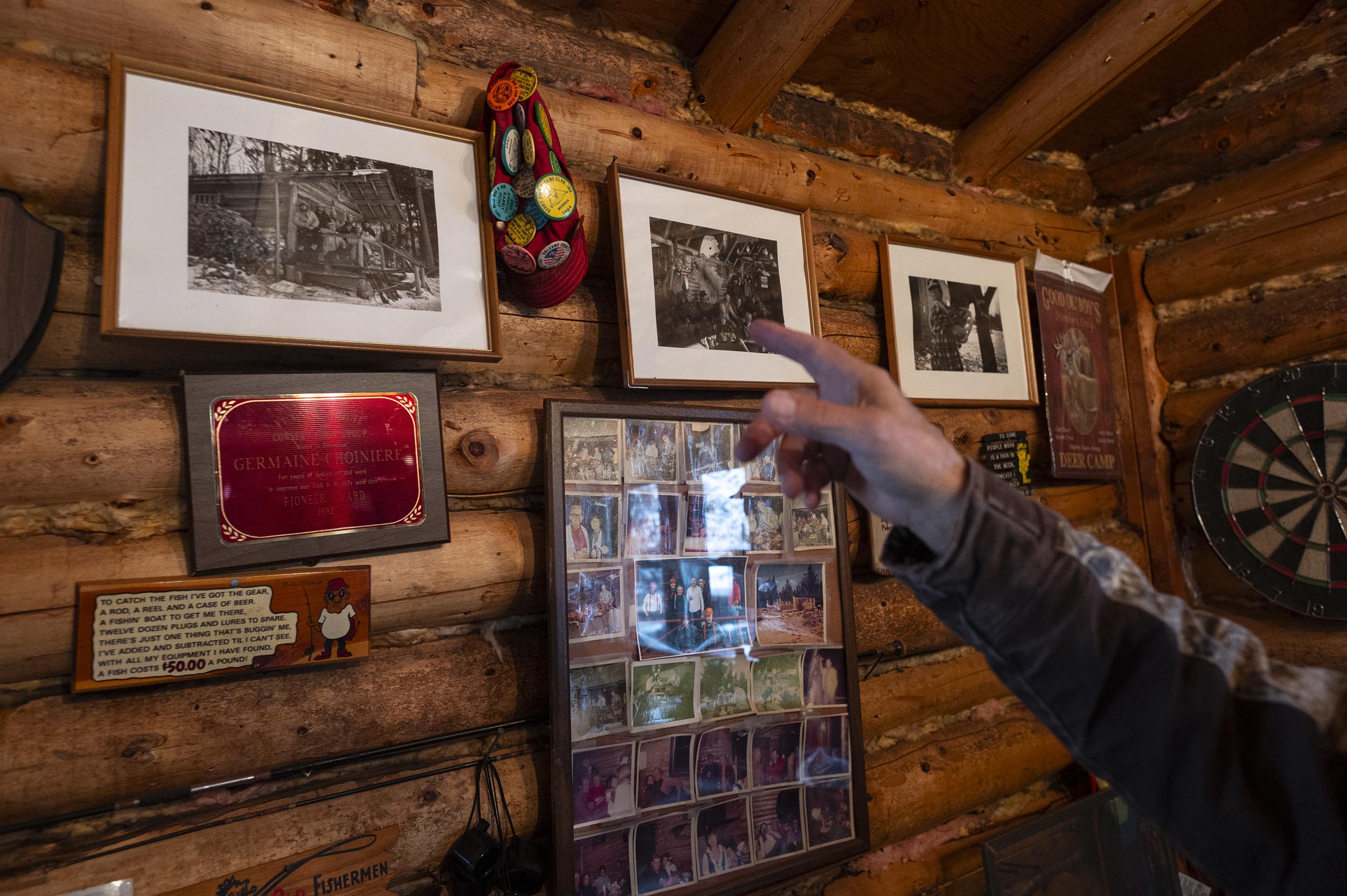 A person points at framed black-and-white photos and a photo collage on a log cabin wall, with a dartboard and various plaques also visible.