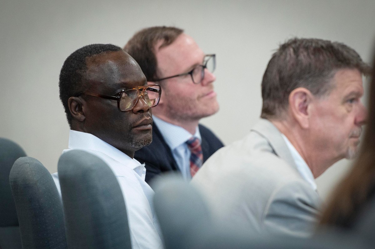 Three men sitting in a row of chairs, facing forward, wearing business attire in what appears to be a formal or professional setting.