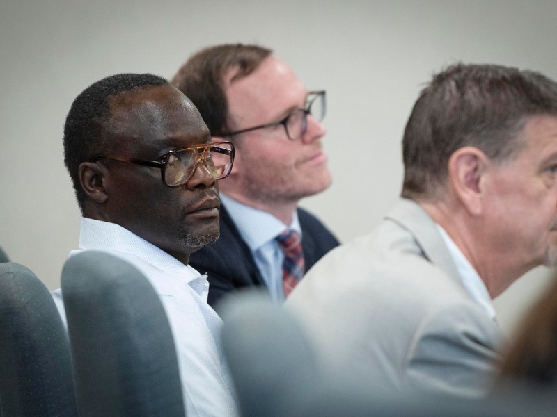 Three men sitting in a row of chairs, facing forward, wearing business attire in what appears to be a formal or professional setting.