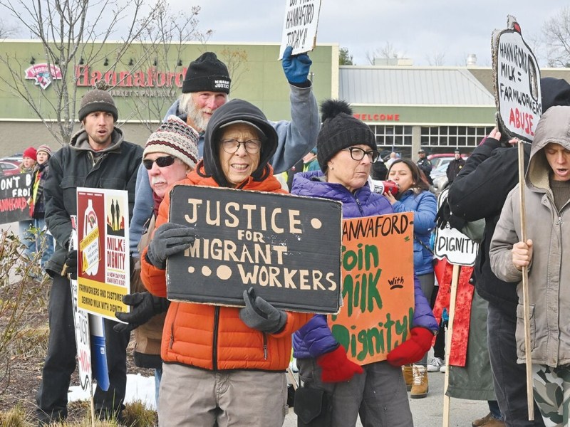 A group of people hold signs advocating for migrant workers' rights outside a Hannaford store, including one sign reading "Justice for Migrant Workers.