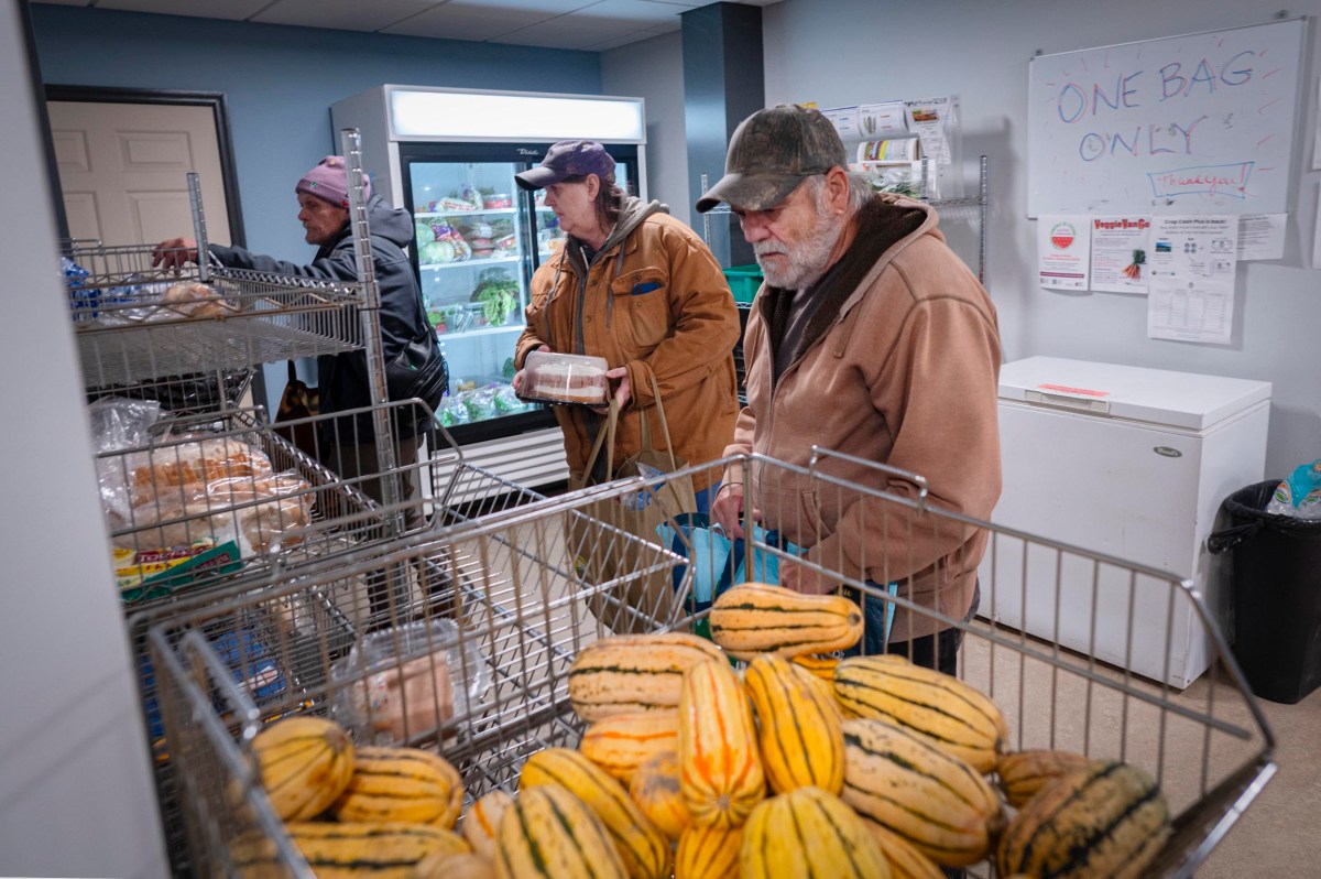 Three people select groceries in a food pantry, with squash and bread in the foreground and a sign reading "One Bag Only" on the wall.