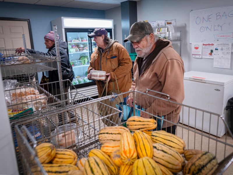 Three people select groceries in a food pantry, with squash and bread in the foreground and a sign reading "One Bag Only" on the wall.