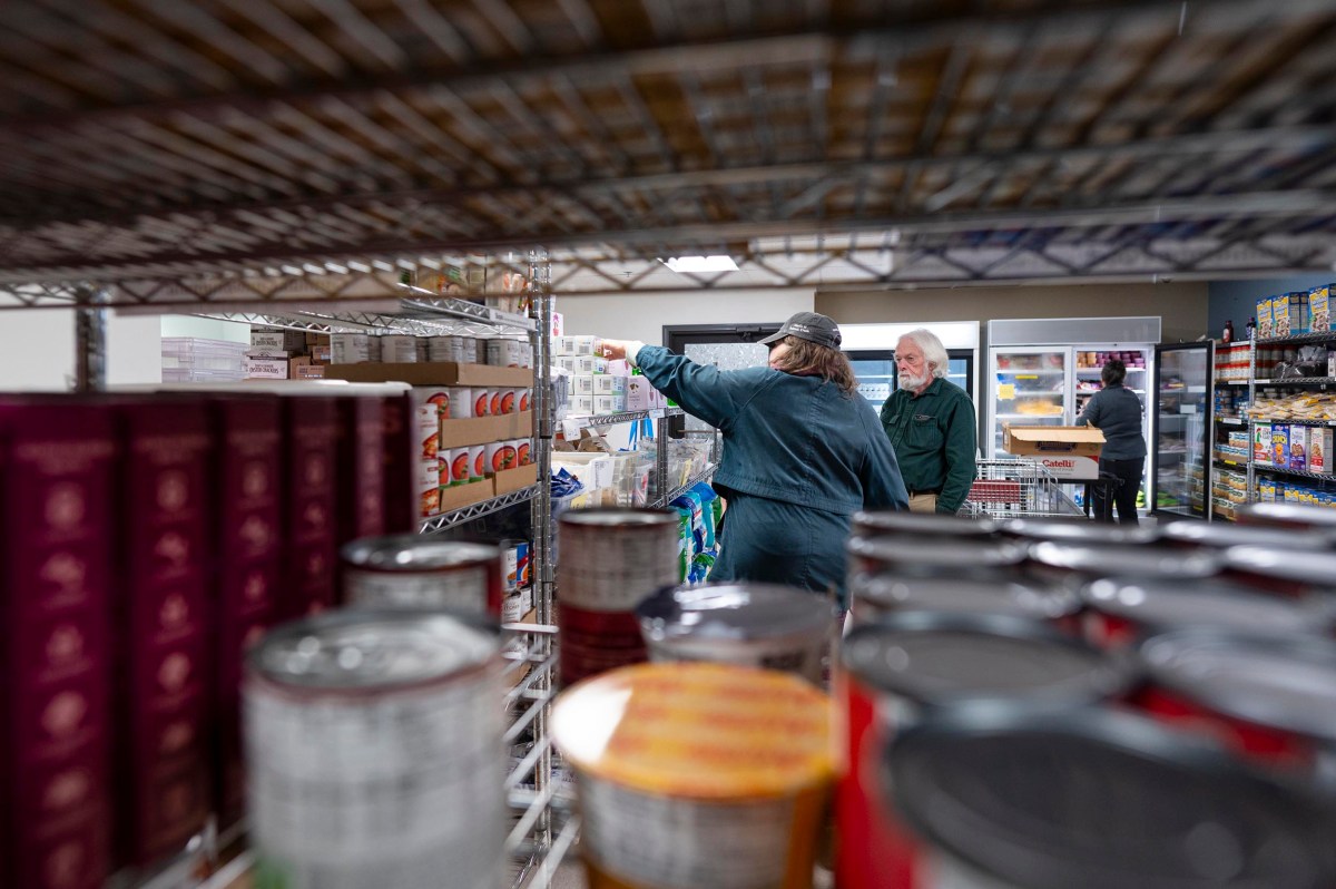 Two people select food items from shelves in a pantry or grocery store, viewed through rows of canned and boxed goods.