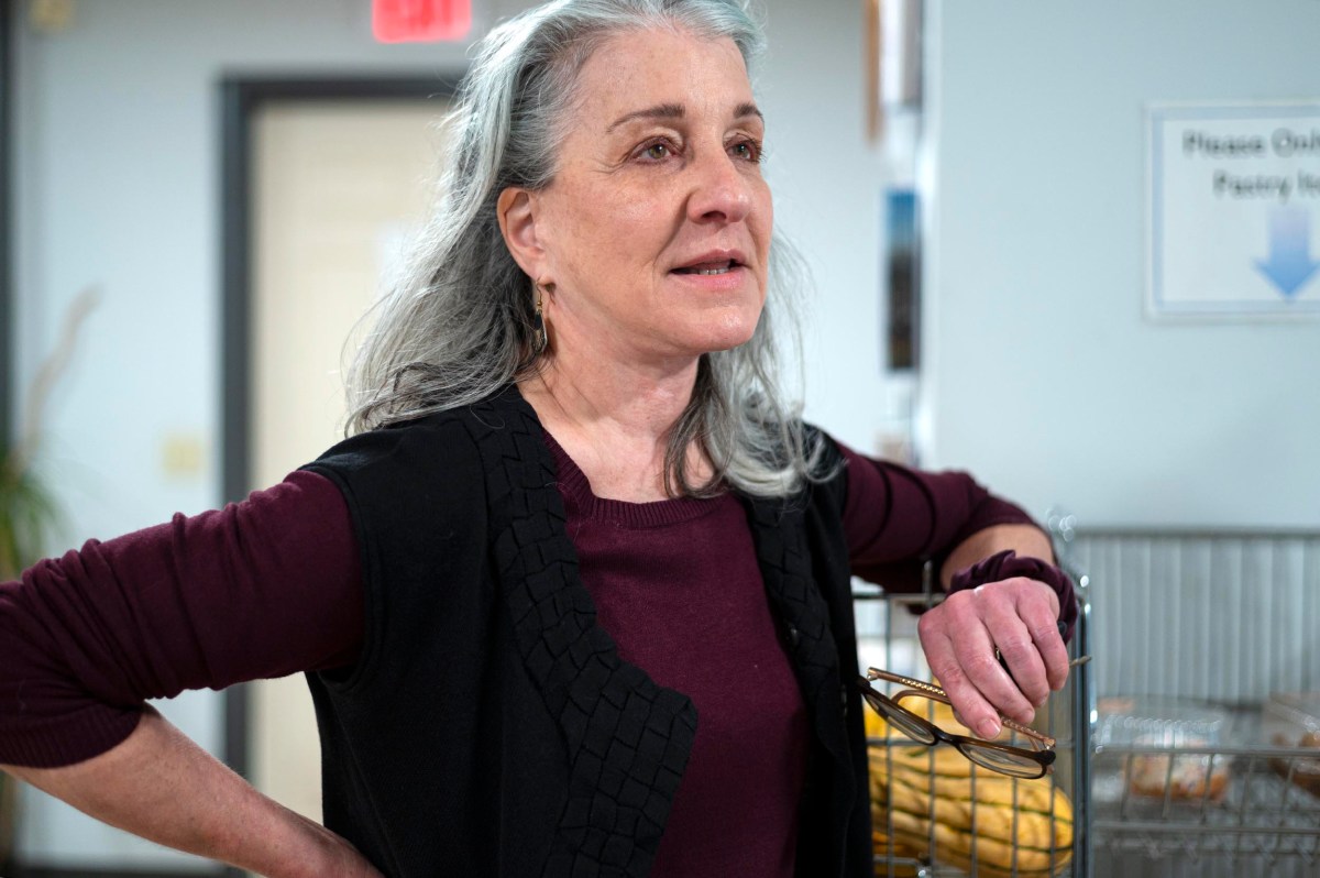A woman with gray hair and a burgundy shirt stands indoors, resting her arm on a cart while holding a pair of eyeglasses.
