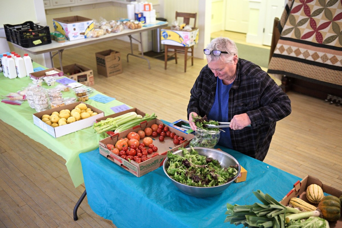 A person prepares fresh salad greens at a table with various fruits and vegetables in a community food distribution setting.