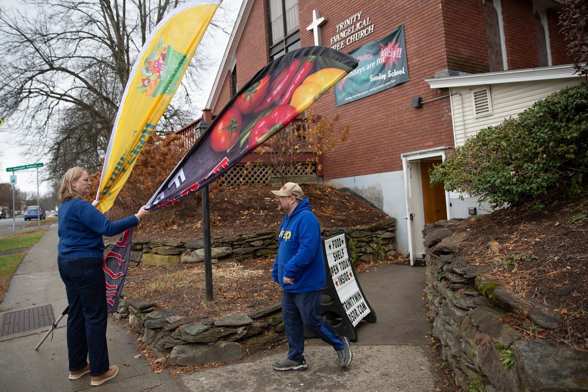 A woman adjusts promotional flags outside a brick church building as a man walks by the entrance on a cloudy day.