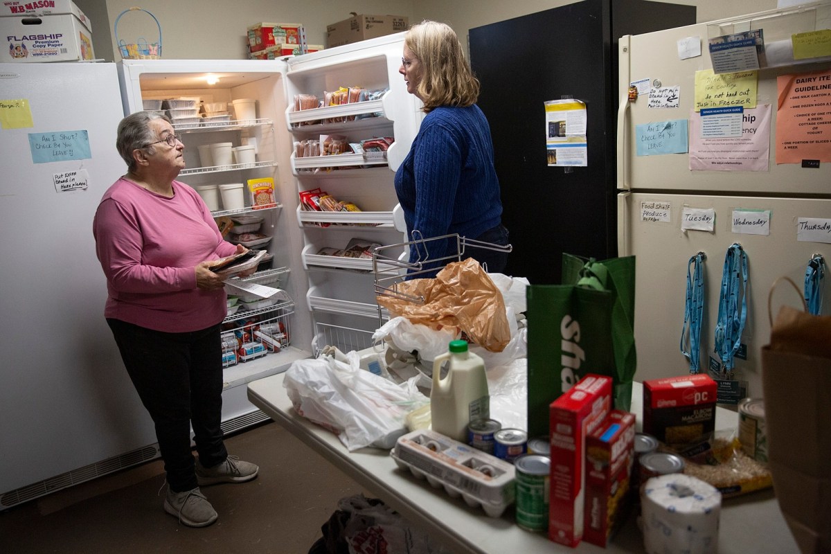 Two women stand in a small room with open refrigerators, surrounded by groceries and food items on a table, appearing to organize or distribute supplies.