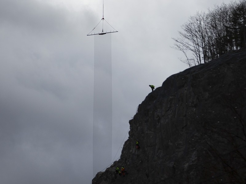 Two workers climb a steep rocky cliff while a large object is suspended by a crane above them against a cloudy sky.