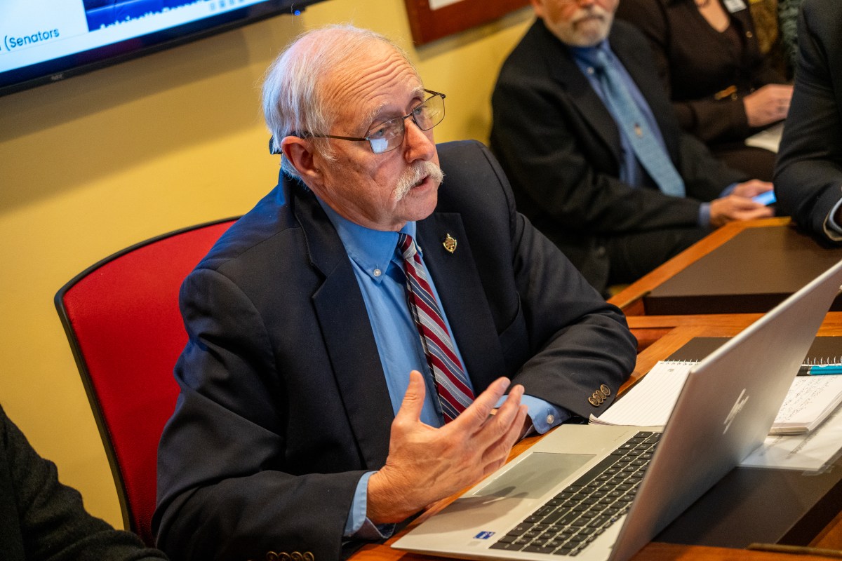 An older man in a suit and tie sits at a desk with a laptop, gesturing as he speaks during a meeting. Other people in business attire sit nearby.