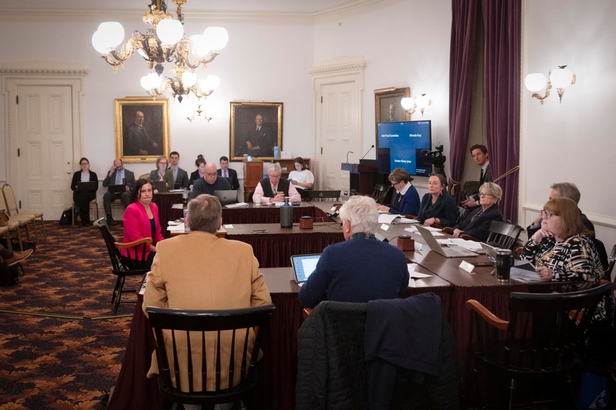 People sit around a large table in a formal meeting room with portraits on the wall and a screen displaying information in the background.