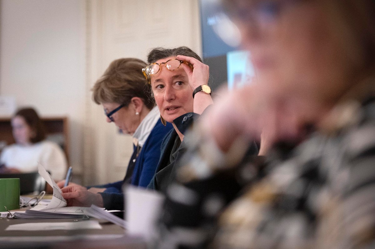 Three women sit at a conference table with papers and pens; one woman in the center adjusts her glasses and looks forward.