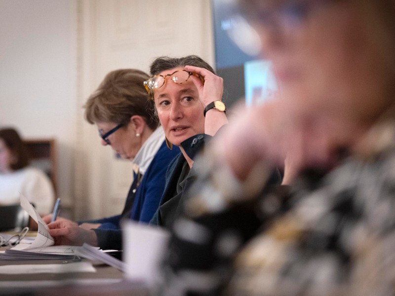 Three women sit at a conference table with papers and pens; one woman in the center adjusts her glasses and looks forward.
