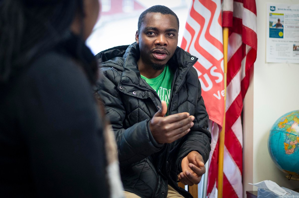 A man wearing a black jacket gestures while speaking to another person in an office with an American flag and a globe in the background.
