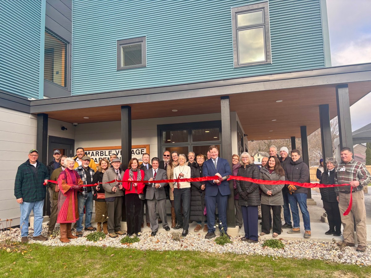 A large group of people stands in front of a modern building, cutting a red ribbon for a ceremony. A sign reads "Warbler Village Apartments.