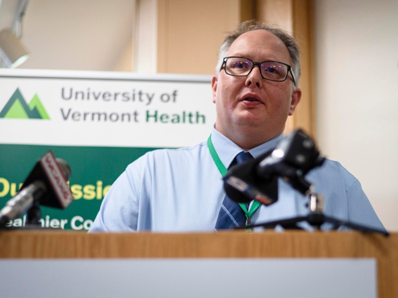 A man in a shirt and tie speaks at a podium with microphones, in front of a University of Vermont Health sign.