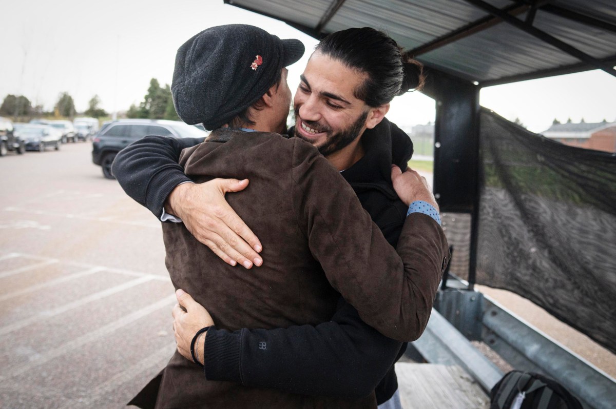 Two men embrace and smile in a parking lot under a metal shelter, with parked cars and trees visible in the background.