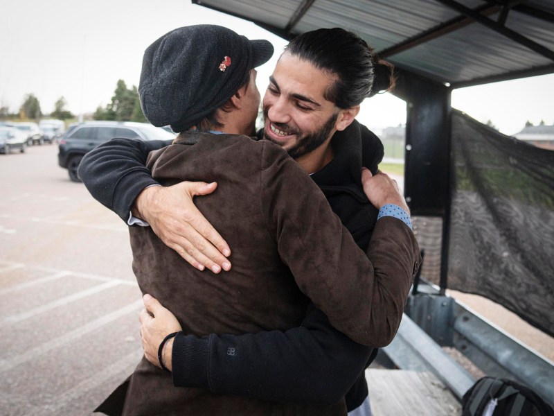 Two men embrace and smile in a parking lot under a metal shelter, with parked cars and trees visible in the background.