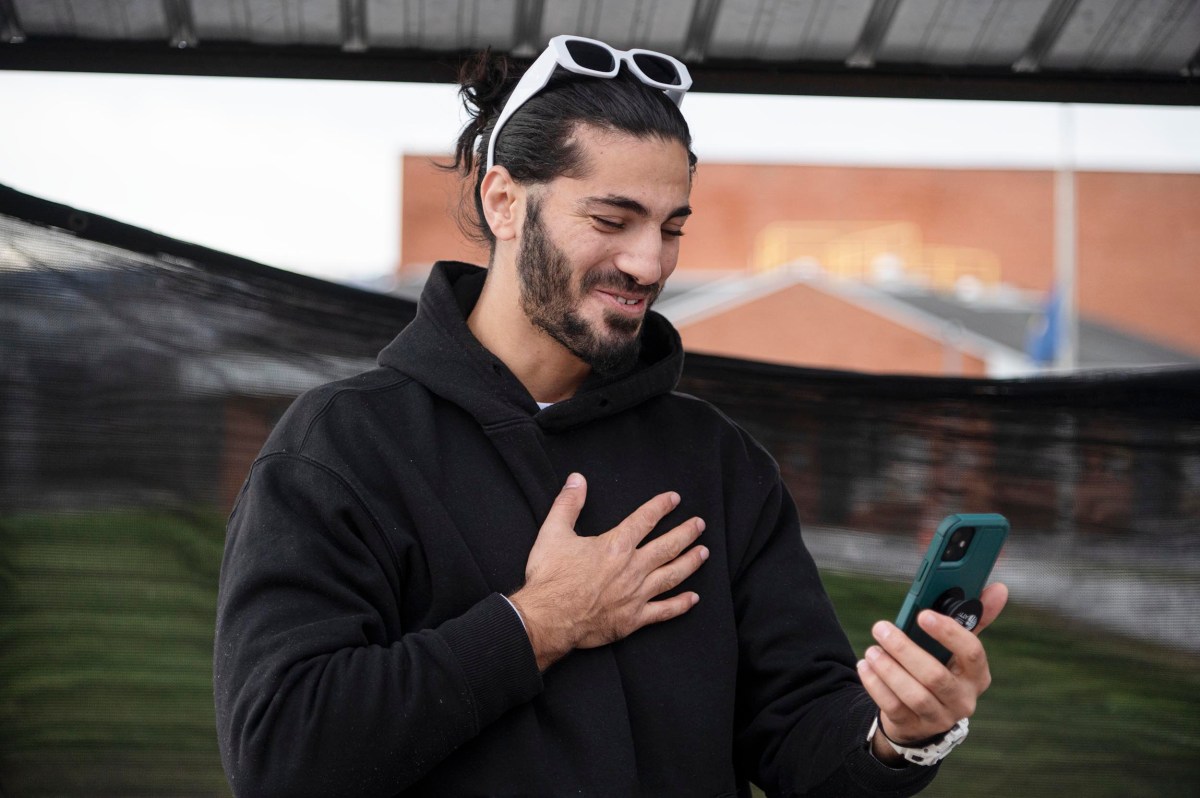 A man with sunglasses on his head smiles and holds a hand to his chest while looking at his phone outdoors.