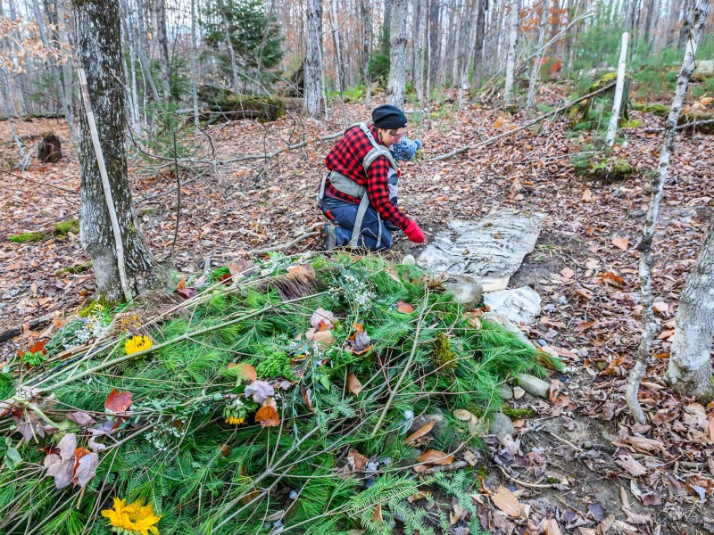 Person in plaid shirt kneels at a grave covered with evergreen branches and flowers in a forest during autumn.