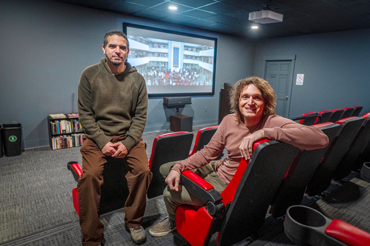 Two people are sitting in a small theater with red and black seats, facing the camera. A screen behind them displays an image of a crowd in a building.