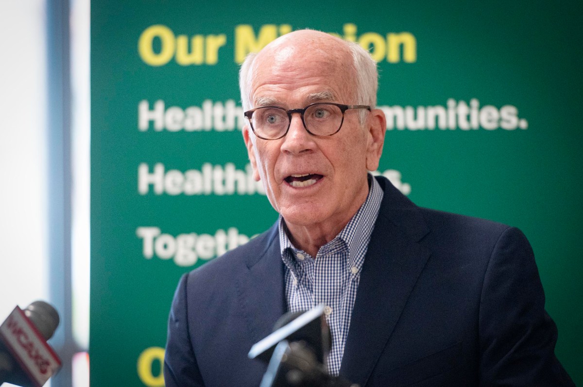 An older man wearing glasses and a suit speaks at a podium, with a green sign about health and community visible in the background.