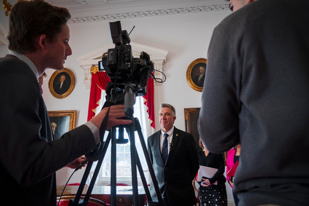 A man in a suit stands in a formal room with portraits on the walls, while two people film him with a video camera.