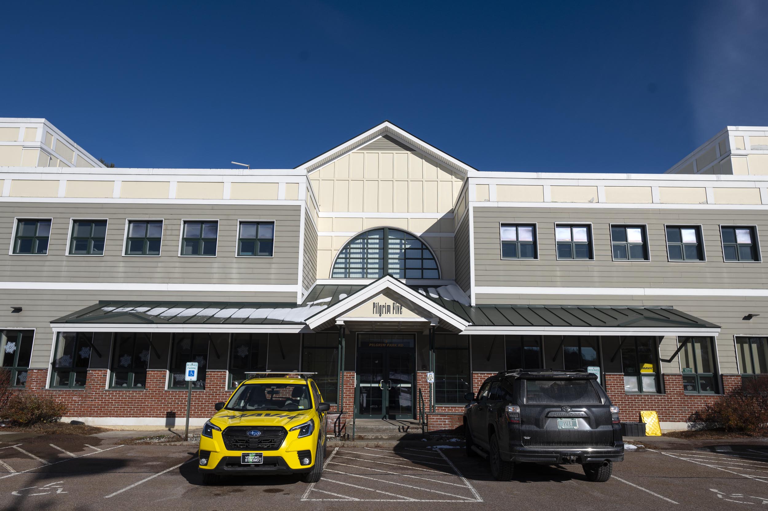 A beige and white two-story building with a peaked roof, large central window, and two cars parked in front, one yellow and one black.
