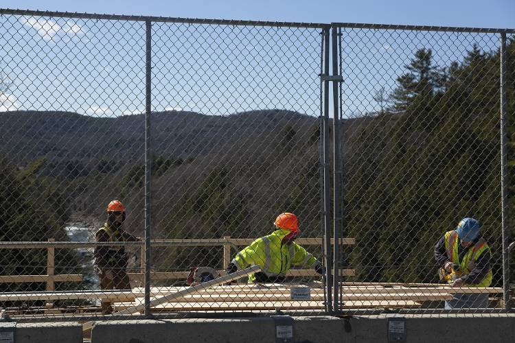Three construction workers wearing safety gear work behind a chain-link fence on a sunny day, with trees and hills visible in the background.