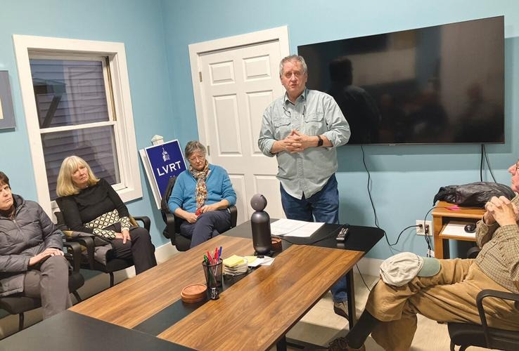 A man stands and speaks to four seated people in a meeting room with blue walls, a large TV, and a table with office supplies.