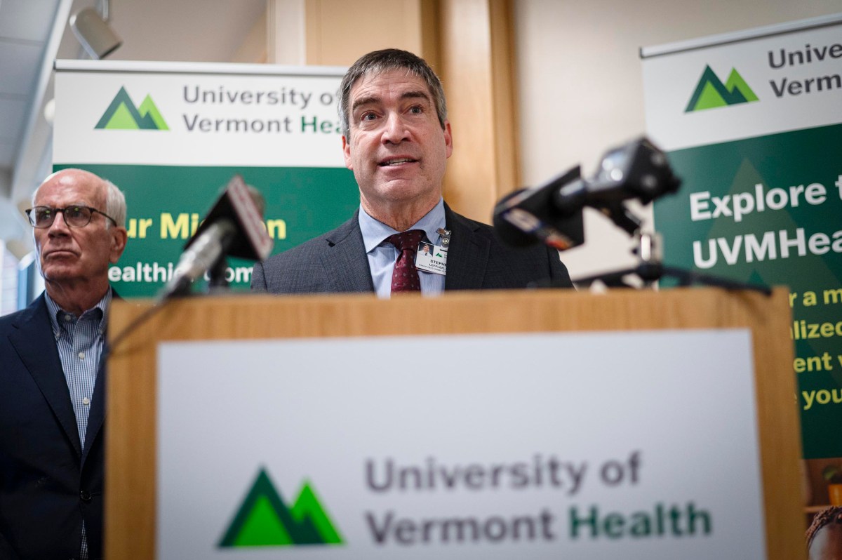 A man speaks at a podium with "University of Vermont Health" signage, while another man stands nearby. Microphones are visible in front of the speaker.
